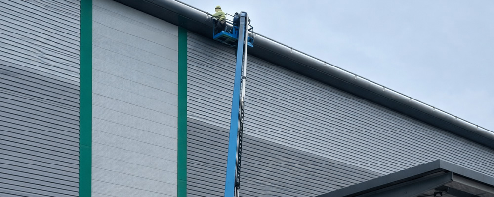 Operative working at height from a mobile boom lift inspecting roof and façade panels on a large industrial warehouse building